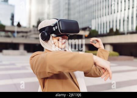 Felice giovane donna d'affari che indossa simulatore di realtà virtuale gesturing al parco d'ufficio in giorno di sole Foto Stock
