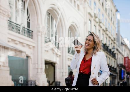 Donna che ride e invia messaggi vocali tramite telefono cellulare in città Foto Stock