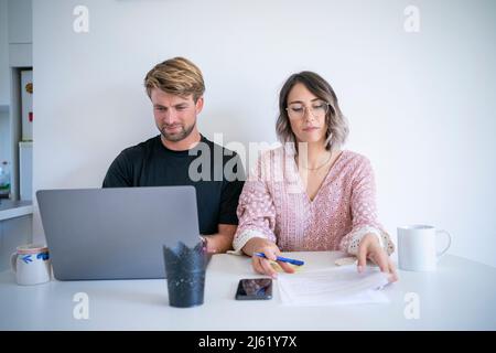 Freelance che legge i documenti da uomo usando il laptop seduto al tavolo da pranzo Foto Stock