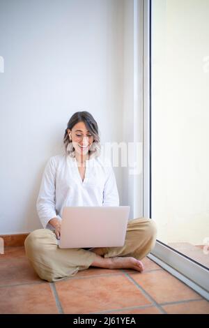 Felice giovane freelance che fa videochiamata attraverso il portatile seduto a gambe incrociate da vetro finestra Foto Stock