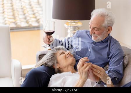 Sorridente donna anziana sdraiata sul braccio del marito seduta con wineglass sul divano a casa Foto Stock