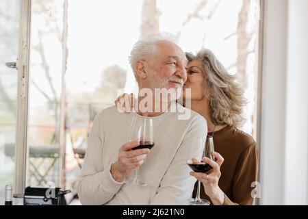 Donna anziana che tiene il wineglass baciare l'uomo di fronte alla finestra dell'appartamento dell'hotel Foto Stock