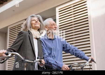 Coppia anziana felice in piedi con biciclette di fronte all'edificio Foto Stock