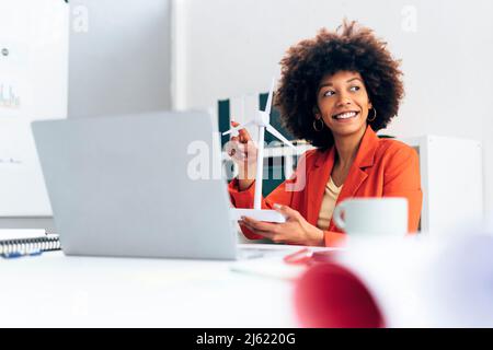 Donna d'affari felice che tiene il modello del mulino a vento seduta con il laptop alla scrivania Foto Stock