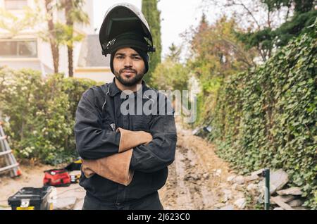 Saldatore che indossa il casco da saldatura in piedi con le braccia incrociate in cantiere Foto Stock