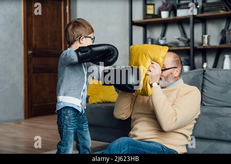 Ragazzo giocoso che indossa guanti da boxe giocando con il viso di copertura del nonno con cuscino nel soggiorno Foto Stock