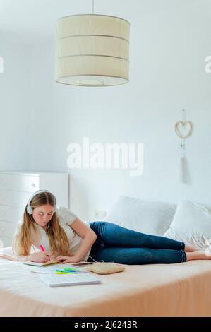 Giovane donna che indossa le cuffie scrivere in blocco appunti sdraiato sul letto a casa Foto Stock