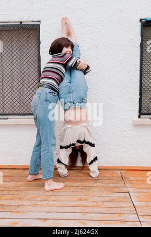 Donna sorridente che aiuta l'amico a fare il cavalletto di fronte al muro bianco sul retro del cortile Foto Stock