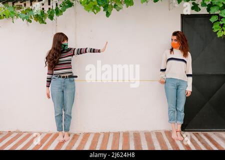 Donna che indossa maschera protettiva che mostra stop gesture a un amico che tiene il nastro di misurazione nel cortile posteriore Foto Stock