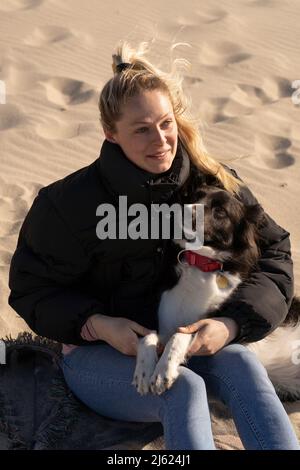 Sorridente giovane donna bionda seduta con cane da compagnia sulla sabbia Foto Stock