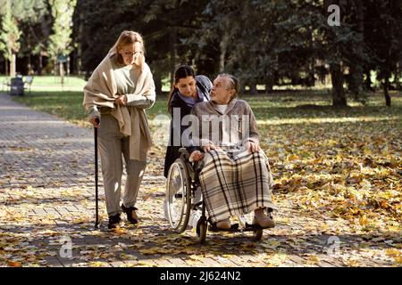 Uomo anziano disabile che parla con la donna che cammina con il bastone da custode che spinge la sedia a rotelle al parco Foto Stock