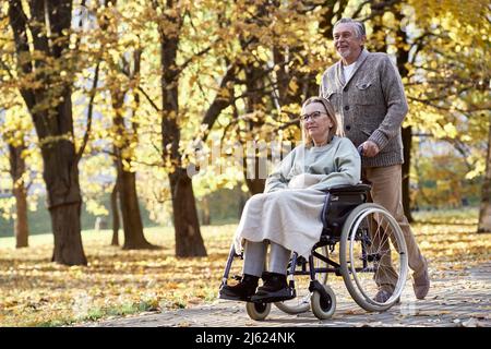 Uomo anziano che cammina con una donna disabile seduta in sedia a rotelle al parco Foto Stock