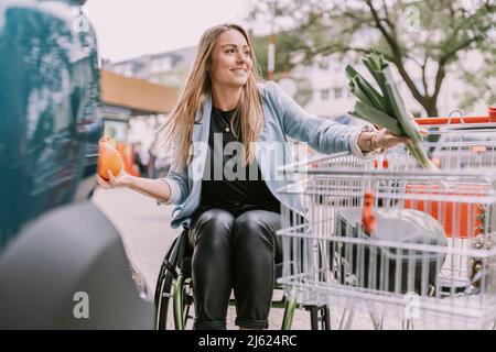 Donna sorridente con generi alimentari seduti in sedia a rotelle da carrello Foto Stock