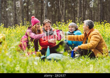 Uomini e donne sorridenti tostano bottiglie e tazze seduti nella foresta Foto Stock