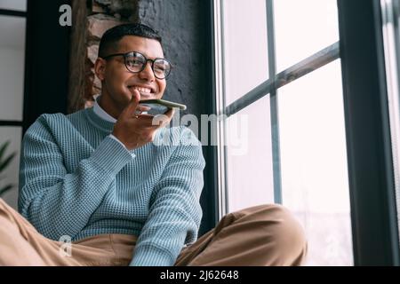 Felice uomo giovane che invia la segreteria telefonica attraverso il telefono cellulare seduto dalla finestra a casa Foto Stock