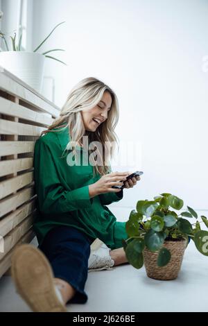 Donna felice usando il telefono intelligente seduto da houseplant a casa Foto Stock