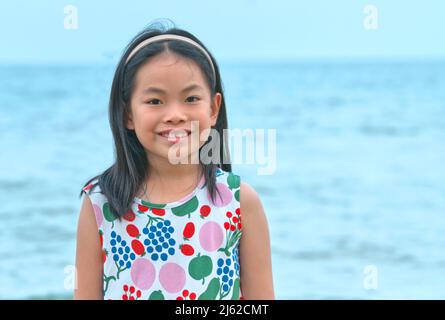 Ritratto di ragazza asiatica, viso sorridente, capelli neri lunghi, sfondo sfocato mare e cielo. Bambina di età compresa tra 8 e 9 anni.
