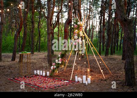 Arco di tibi bohémien fatto di aste di legno decorate con rose rosa, candele su tappeto, erba pampass, avvolto in luci fata su matrimonio all'aperto Foto Stock