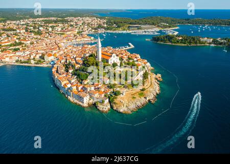 Il motoscafo che naviga intorno a Rovigno. Dintorni di Rovigno e torre della chiesa di Sant'Eufemia. Edifici della città croata vicino alle foreste e al mare Adrianico alla luce del sole. Panorama aereo Foto Stock