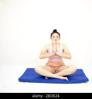bella donna in posa yoga con le mani rialzate per pregare e occhi chiusi su tappeto blu con sfondo bianco Foto Stock