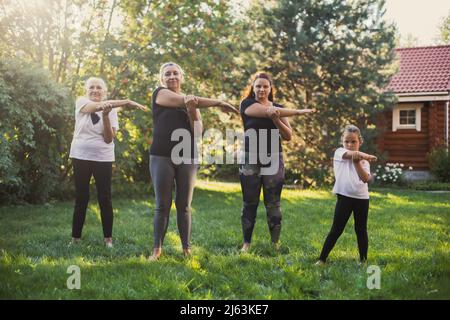 Femmine di poche generazioni che si esercitano all'esterno allungando le braccia insieme nello stesso modo in piedi su prato pieno di erba verde e alberi. Trascorrere del tempo Foto Stock