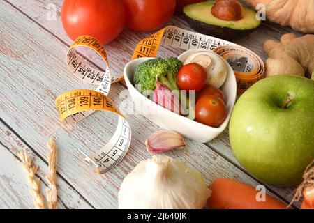 Concetto sano di controllo del peso da mangiare con frutta e verdura in una ciotola a forma di cuore e intorno e nastro di misurazione sul dettaglio del legno. Vista rialzata. Hor Foto Stock