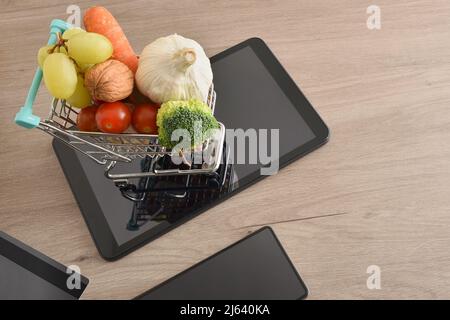 Shopping online background con dispositivi mobili e mini supermercato carrello pieno di frutta e verdura su tavola di legno. Vista dall'alto. Orizzontale Foto Stock