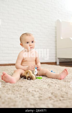 il bambino cute siede su un tappeto in un pannolino usa e getta gioca con i giocattoli contro una parete di mattone bianca. Foto Stock