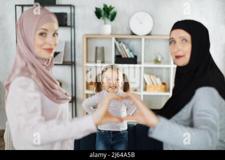 Ritratto di felice tre generazioni di donne islamiche posa insieme mostra amore cuore gesto mano. Sorridendo bambina musulmana con giovane madre e nonna anziana si sentono uniti. Foto Stock