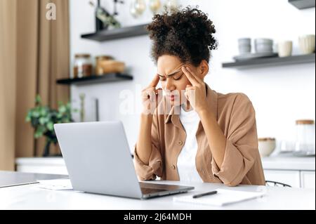 Overworking frustrato triste african american ragazza, studente o freelancer, in abiti casual, studiare o lavorare a casa utilizza computer portatile, vivendo stress, la sofferenza di mal di testa, emicrania, bisogno di riposo Foto Stock