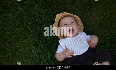 Piccolo bambino neonato felice in estate panama cappello cadere giù posa su erba a piedi nudi in estate Sunny Day. Bambino bambino bambino ragazzo Smacinatura faccia guardare Foto Stock