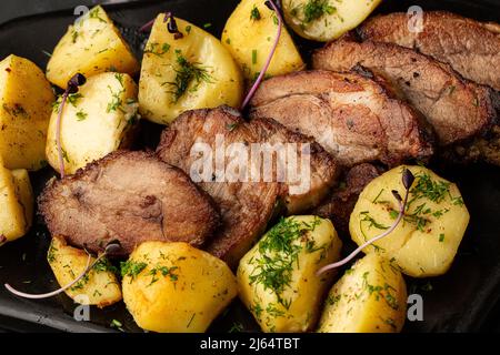 Patate novelle al forno con sale marino e filetto di maiale tagliato a pezzi in un piatto nero, su tavola di marmo Foto Stock