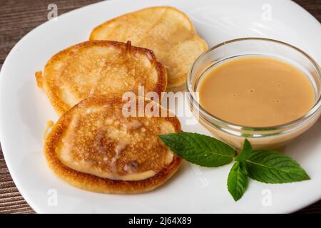 Mucchio di frittelle spesse fritte di fresco, nelle cucine dell'Europa orientale chiamate oladky o oladyi con latte condensato su sfondo di legno. Foto Stock