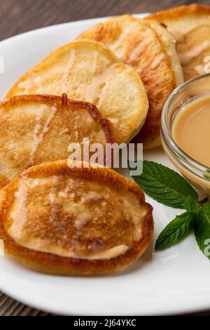 Mucchio di frittelle spesse fritte di fresco, nelle cucine dell'Europa orientale chiamate oladky o oladyi con latte condensato su sfondo di legno. Foto Stock