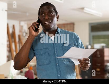 Perplesso africano americano con carte che parlano al telefono durante la ristrutturazione della casa Foto Stock
