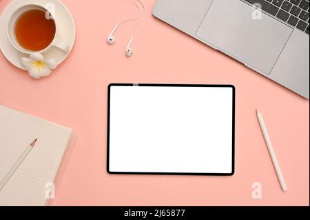 Splendida scrivania da lavoro femminile con vista dall'alto, computer portatile, teacup, cancelleria e tablet digitale, schermo bianco su sfondo rosa. Foto Stock