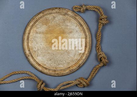 Tavola rotonda di taglio in legno e corda della nave sullo sfondo grigio. Svuotare il vecchio tagliere. Corda doppia di iuta con nodi semplici legati. Vista dall'alto. Foto Stock