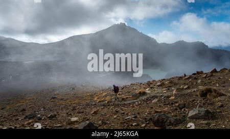 Persone che camminano lungo il ripido terreno di scrosco dal Red Crater ai laghi di smeraldo sul Tongariro Alpine Crossing. Fumetti vulcanici che si sgocciolano sulla valle, N Foto Stock