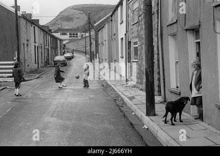 Bambini che giocano a calcio in strada e una signora sul suo ingresso, Long Row, Blaenlechau, Rhondda Fach, Galles del Sud, 1984 Foto Stock