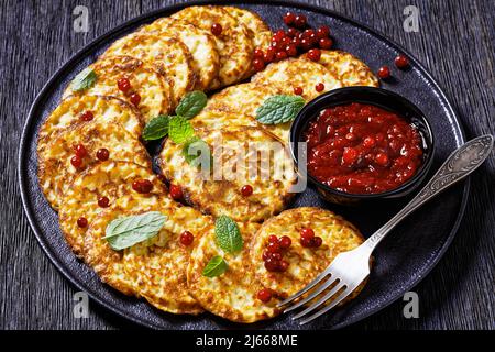 frittelle dolci di farine di farro con salsa di bacca, mirtillo di montagna, cowberry su piatto nero su tavola di legno scuro, vista orizzontale dall'alto, primo piano Foto Stock