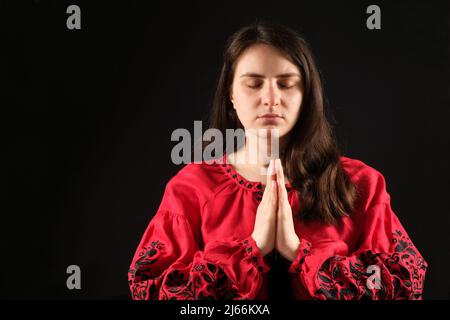 Una giovane donna con gli occhi chiusi in una camicia rossa ha piegato le mani insieme in preghiera, uno sfondo nero, un luogo per il testo. Foto Stock