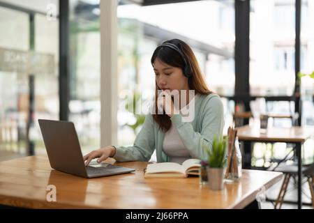 giovane donna asiatica indossare cuffie studiare online guardare podcast webinar su laptop ascoltare apprendimento corso di formazione conference call prendere appunti Foto Stock