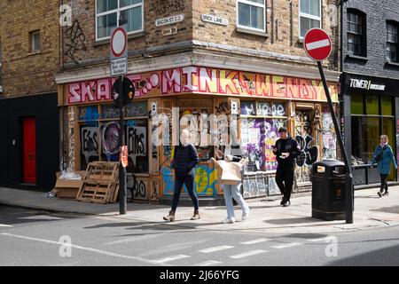 Persone che guardano i cellulari che camminano lungo Brick Lane da Bacon Street passato un negozio chiuso coperto di graffiti a East London E1 Inghilterra Regno Unito KATHY DEWITT Foto Stock