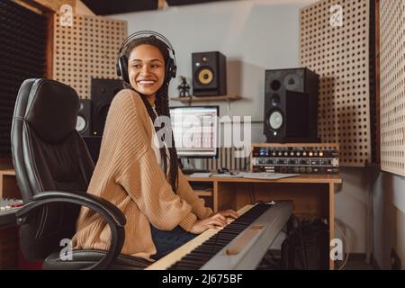 Attraente musicista musicale femminile afroamericana di stile che suona sintetizzatore mentre lavora in studio di registrazione Foto Stock