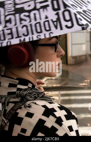 Belgrado, Serbia - 27 aprile 2022: Giovane donna in abito bianco e nero con cuffie rosse, rossetto rosso e occhiali sotto ombrello Foto Stock