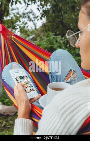 Donna blogger leggere il commento su smartphone dispositivo bere tè relax in amaca in autunno estate primavera.Donna guardando il cellulare schermo di navigazione internet Foto Stock