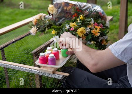 Arte e mestieri adulti concetto di creatività terapia d'arte, l'uomo dipinge fiori di seta in ambiente tranquillo all'aperto con sfondo verde erba e stampelle appoggiate Foto Stock