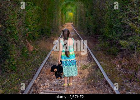 Bambina in dietro, vestita con un lungo vestito che tiene un orsacchiotto in piedi sulle vecchie rotaie in un labirinto di tunnel ferroviario abbandonato. Foto Stock