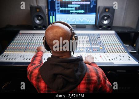 Ripresa posteriore di un tecnico del suono nero irriconoscibile che produce registrazioni in studio regolando le impostazioni audio Foto Stock