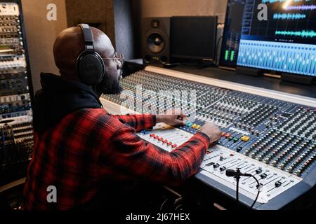 L'elegante uomo afro-americano indossa una camicia rossa e cuffie che lavorano su una traccia musicale in studio di registrazione Foto Stock